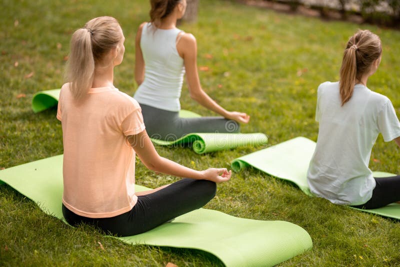Young slim girl sits relaxing in the lotus position doing exercises on yoga mats with other girls on green grass in the royalty free stock image Young slim girl sits relaxing in the lotus position doing exercises on yoga mats with other girls on green grass in the royalty free stock image