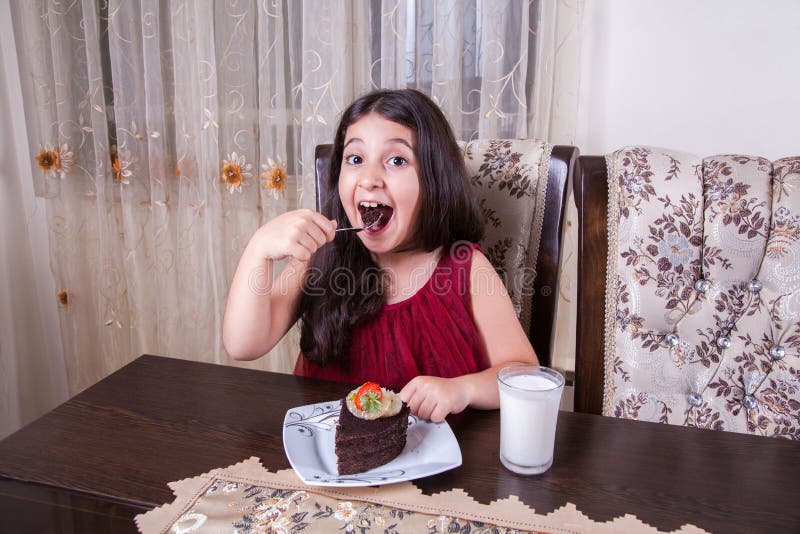 Young small beautiful middle eastern child girl with chocolate cake with pineapple, strawberry, and milk with red dress and dark