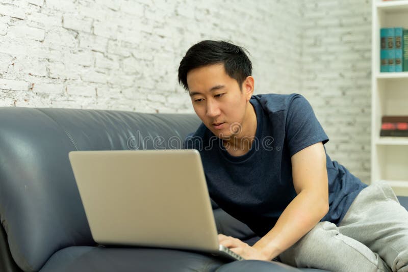 Young smiling Asian man in casual clothing on sofa working on laptop computer. Male freelancer working from home on royalty free stock image Young smiling Asian man in casual clothing on sofa working on laptop computer. Male freelancer working from home on royalty free stock image