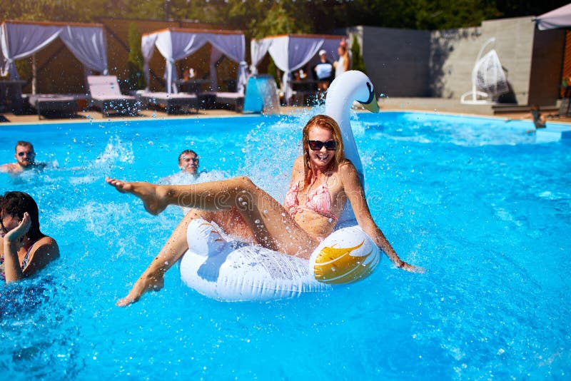 Young smiling bikini girl having fun with friends on inflatable swan in swimming pool. Happy people splashing water on