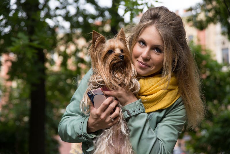 Young smiling blonde woman in city park. Small yorkshire terrier is on her hands.