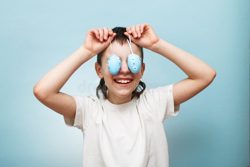 Young smiling boy holding Easter eggs in his hand and covering his eyes with it on a blue background royalty free stock photo