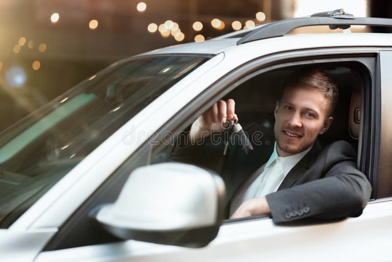 Young smiling businessman sitts in his car looks happy and satisfied, holding car keys, successful business strategy concept.