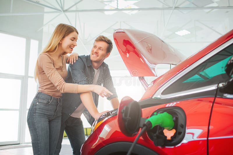 Young smiling family buying first electric car in the showroom. Attractive pleasantly surprised woman point at eco stock photography Young smiling family buying first electric car in the showroom. Attractive pleasantly surprised woman point at eco stock photography