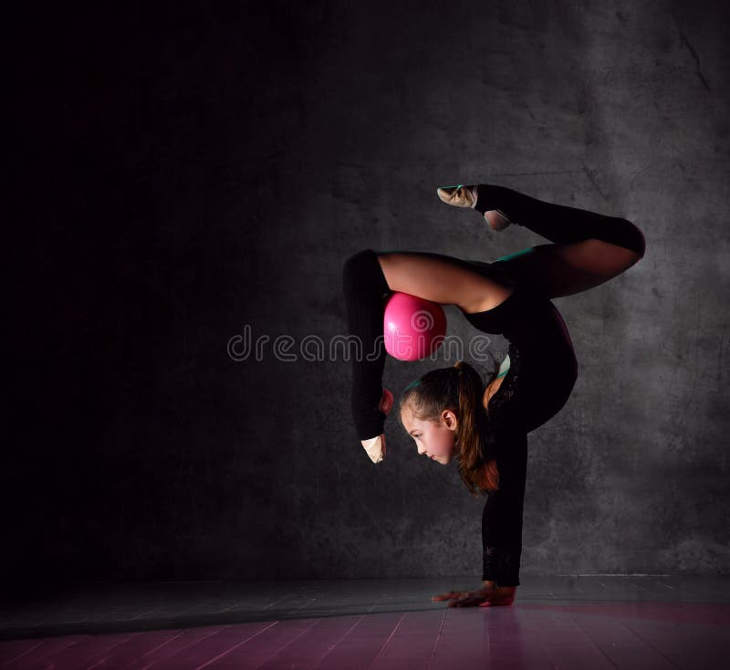 Young girl gymnast in black sport body and uppers standing in bridge pose and holding pink gymnastic ball between leg and neck