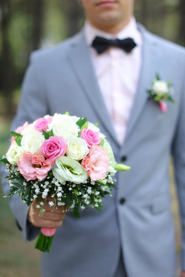 Young smiling groom wearing grey suit keeping bouquet of flowers.