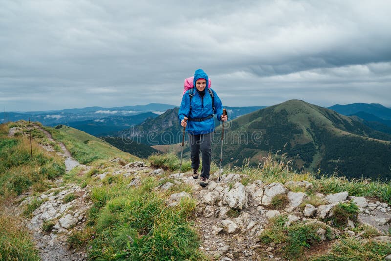 Mladá usmievavá turistka oblečená do dažďa pri prechádzke skalnatým pohorím slovenskej malej fatry pomocou sledovacích palíc s