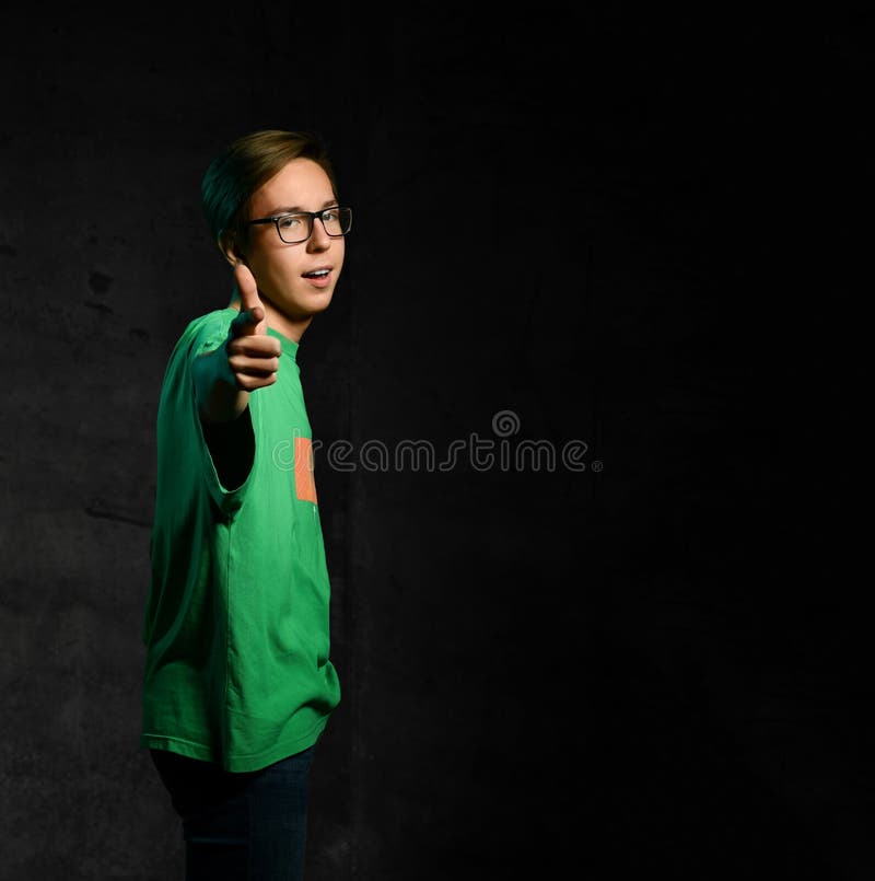 Young smiling teen boy in green t-shirt, jeans and glasses standing and pointing at camera over dark background