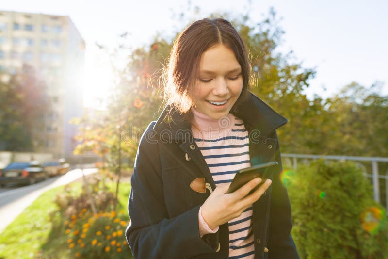 Young smiling teen girl with mobile phone, sunny autumn day, girl in coat, golden hour