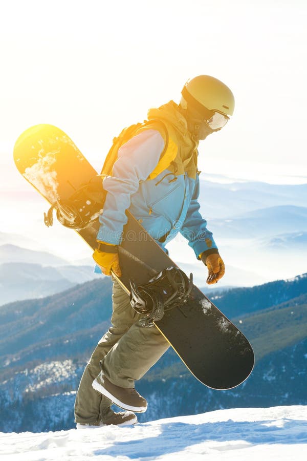 Young snowboarder walking at the top of a mountain with sun behind him