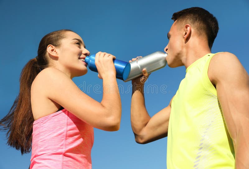 Young sporty couple drinking from water bottles against blue sky