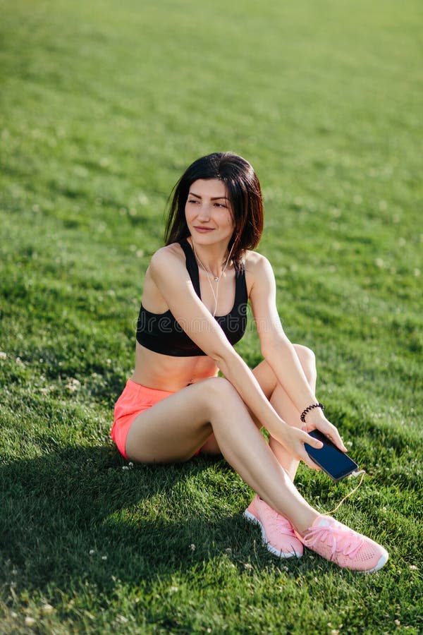 Young sporty joyful woman brunette in shorts and top sitting on the grass football field stadium and listens to music in earphones royalty free stock photo Young sporty joyful woman brunette in shorts and top sitting on the grass football field stadium and listens to music in earphones royalty free stock photo