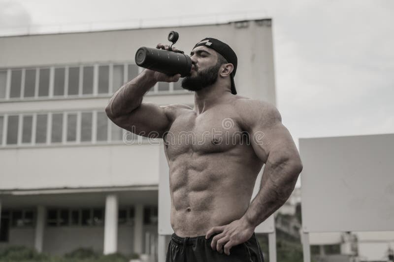 Young strong athlete male drinking water from black bottle during outdoor fitness workout on university building backgroung