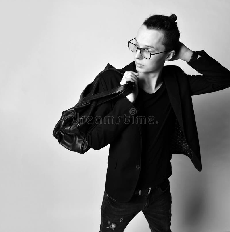 Young stylish man with a bun on head in glasses, black jeans, t-shirt and with a big stylish bag on his shoulder looks back stock photography