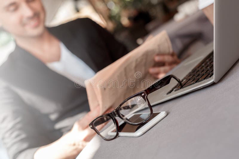 Lunch time. Young man in suit sitting at cafe on city street reading newspaper joyful blurred eyeglasses and phone close stock photography Lunch time. Young man in suit sitting at cafe on city street reading newspaper joyful blurred eyeglasses and phone close stock photography