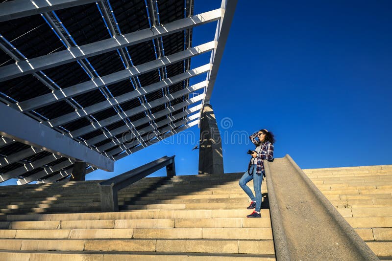 Young and stylish woman is standing on stairs next to the huge solar panel in Port Forum, Barcelona, Spain.
