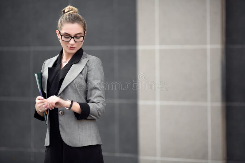 Young successful woman in a strict suit looks at the wristwatch. A woman in the city is holding a file. royalty free stock photography Young successful woman in a strict suit looks at the wristwatch. A woman in the city is holding a file. royalty free stock photography