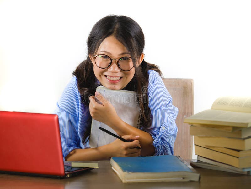 Young sweet and happy Asian Chinese student girl in nerd glasses working cheerful on laptop computer on desk with pile of books