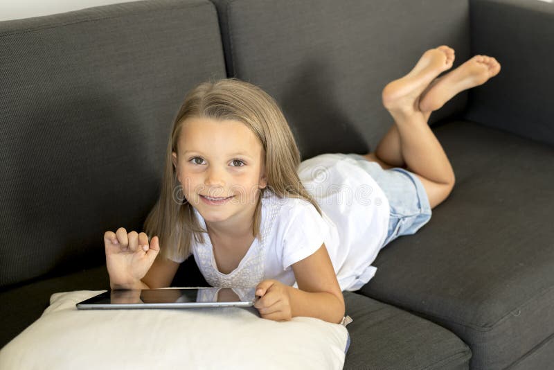 Young sweet and happy little girl 6 or 7 years old lying on home living room sofa couch using internet digital tablet touch pad