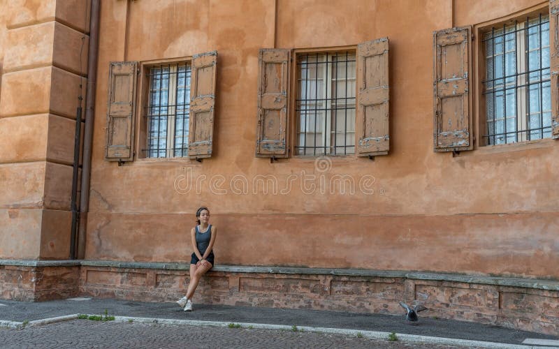 Young teen girl with headband, shorts, tanktop and sneakers sitting on brick bench under 3 windows with shutters and bars