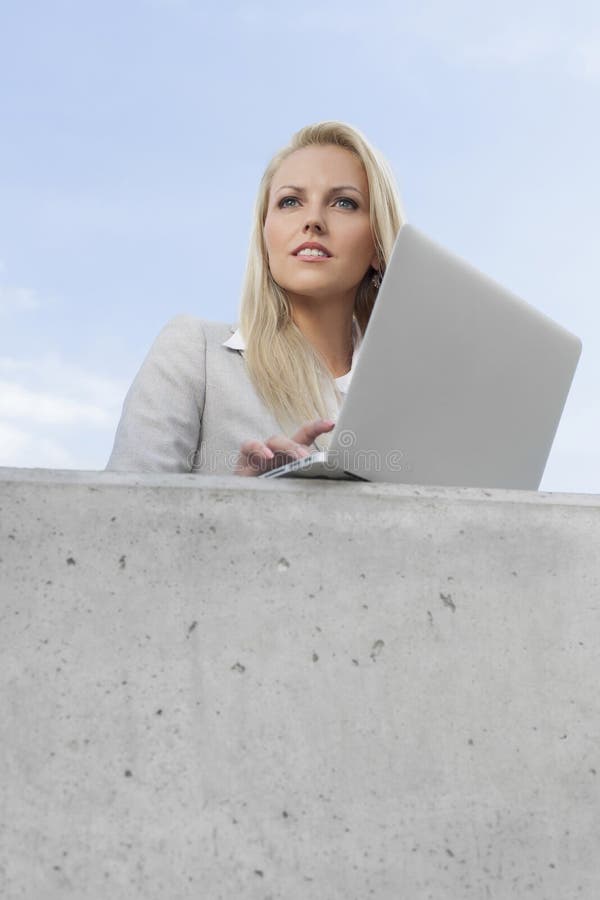 Young thoughtful businesswoman with laptop looking away while standing on terrace against sky