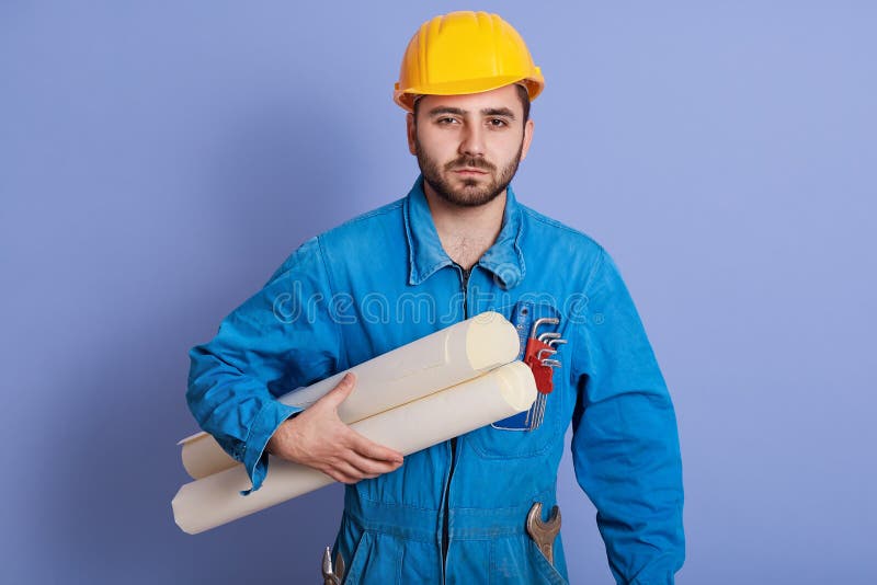 Young thoughtful foreman wearing blue uniform and yellow helmet holding plan of new project in hands, posing with blueprints stock photography Young thoughtful foreman wearing blue uniform and yellow helmet holding plan of new project in hands, posing with blueprints stock photography
