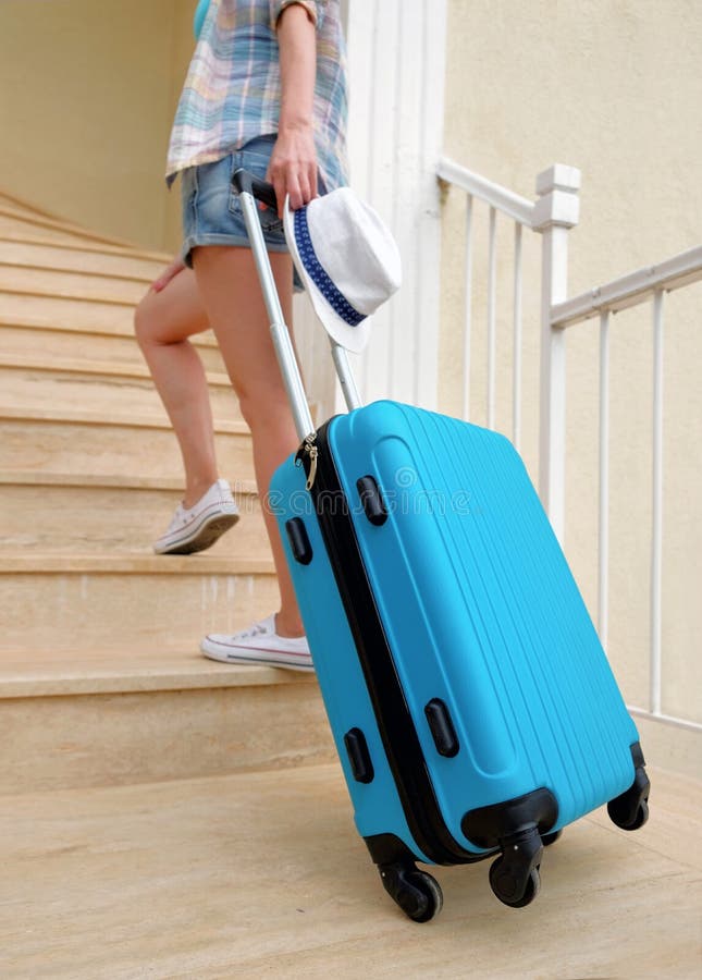 A young travel woman in white sneakers carries a blue suitcase up the stairs to the hotel