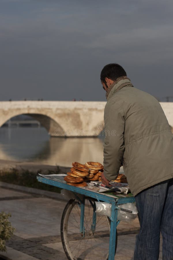 A young man is selling simit Turkish sesame bagel on a wooden food cart in the street. Historic Stone bridge over Seyhan river in Adana is in the background. A young man is selling simit Turkish sesame bagel on a wooden food cart in the street. Historic Stone bridge over Seyhan river in Adana is in the background