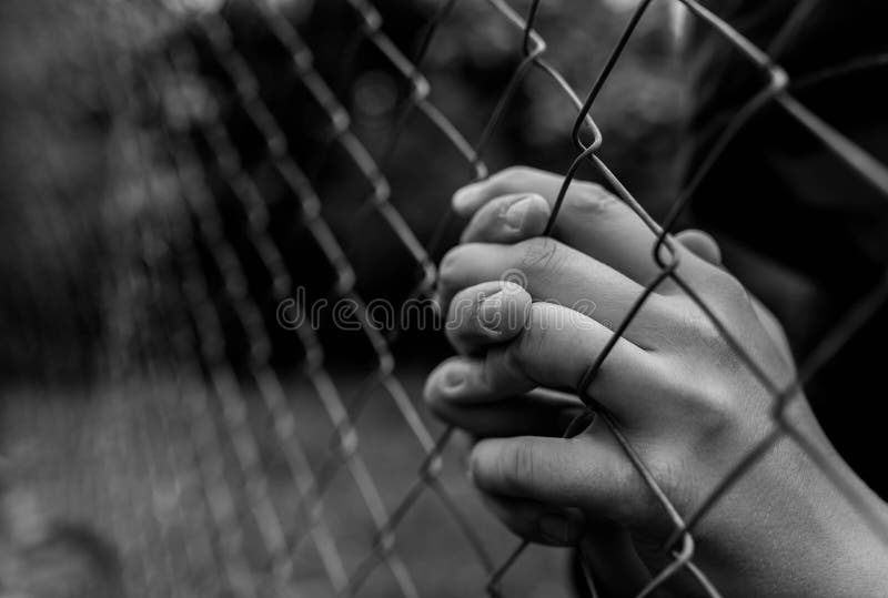 Young unidentifiable teenage boy holding the wired garden  praying at the correctional institute in black and white, conceptual image of juvenile delinquency, focus on the boys hand.