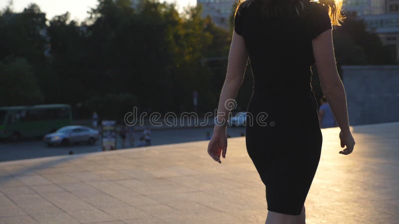 Young unrecognisable girl in black dress walking along urban street at sunset time. Attractive business woman going in