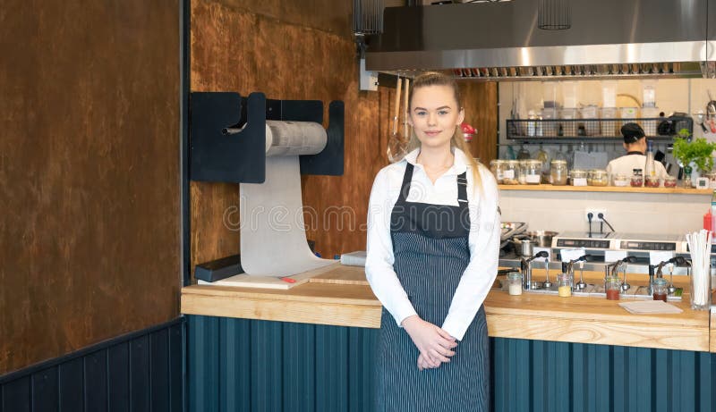 Young waitress wearing apron posing in restaurant. Portrait of a young woman on her first day job in a coffee shop. Smiling young royalty free stock image Young waitress wearing apron posing in restaurant. Portrait of a young woman on her first day job in a coffee shop. Smiling young royalty free stock image