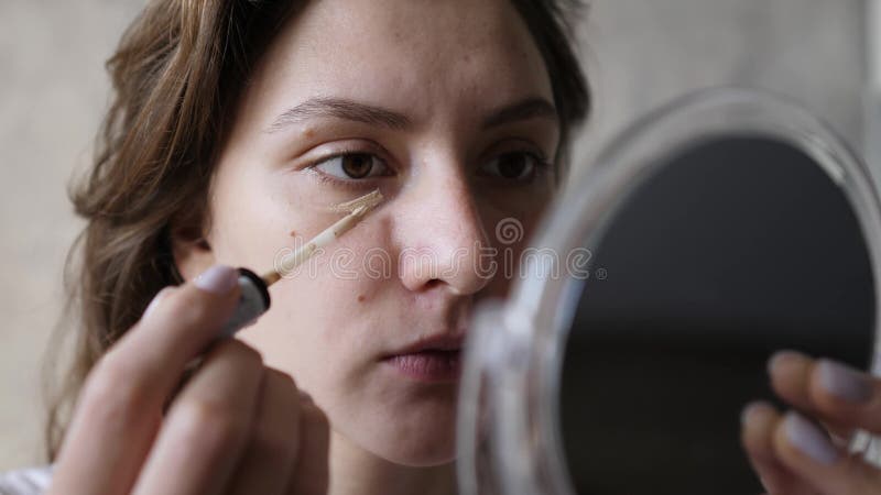 A young woman applies a smear of concealer with a brush under her eye and looks in a small mirror. Basic makeup