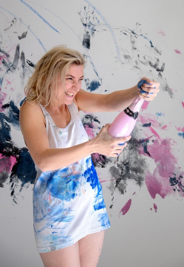 Young woman artist opens a sparkling bottle of champagne in white undershirt in front o the painted wall for her birthday party stock image Young woman artist opens a sparkling bottle of champagne in white undershirt in front o the painted wall for her birthday party stock image