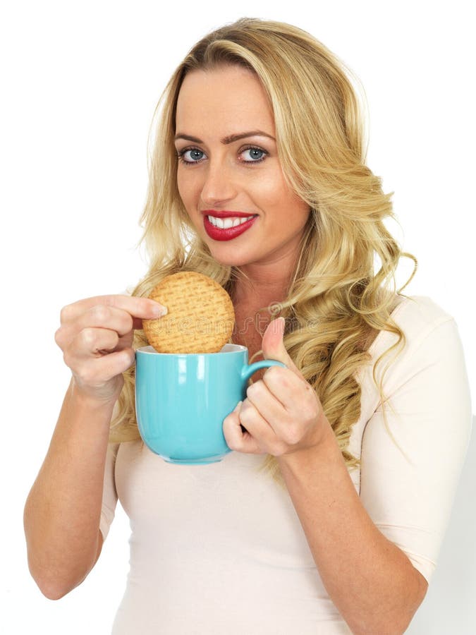 Attractive Woman, with long blonde hair and in her twenties, holding a blue mug of tea or coffee and dunking a biscuit in the mug. Taking a break or having elevenses, looking at the camera, happy and smiling, wearing a cream top and red lipstick. Shot against a white background. Attractive Woman, with long blonde hair and in her twenties, holding a blue mug of tea or coffee and dunking a biscuit in the mug. Taking a break or having elevenses, looking at the camera, happy and smiling, wearing a cream top and red lipstick. Shot against a white background.