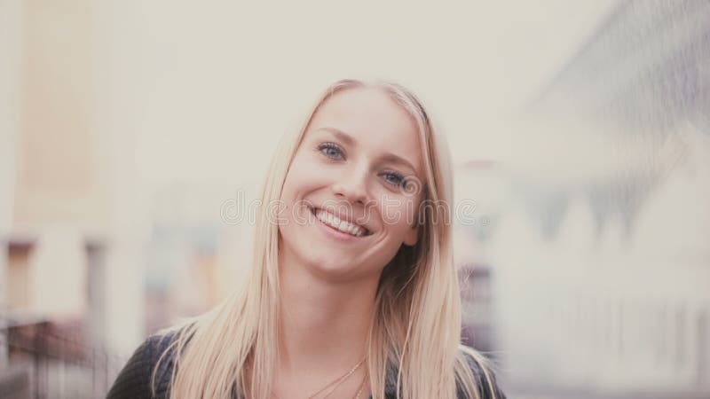 Young woman with blonde long hair looking at camera and smiling. Portrait of attractive girl in city blur background.