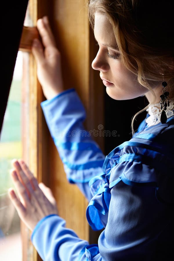 Young woman in blue vintage dress standing near window in coupe stock photography Young woman in blue vintage dress standing near window in coupe stock photography