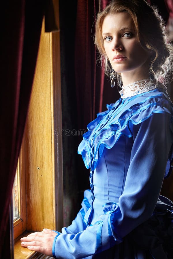 Young woman in blue vintage dress standing near window in coupe stock images Young woman in blue vintage dress standing near window in coupe stock images