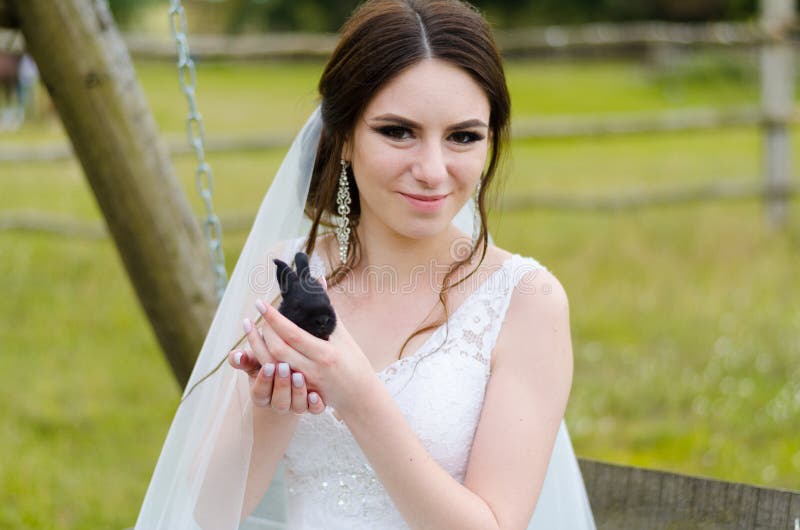 Young woman bride smiling and holding cute rabbit over park summer nature outdoor. White wedding dress, green background royalty free stock photography Young woman bride smiling and holding cute rabbit over park summer nature outdoor. White wedding dress, green background royalty free stock photography