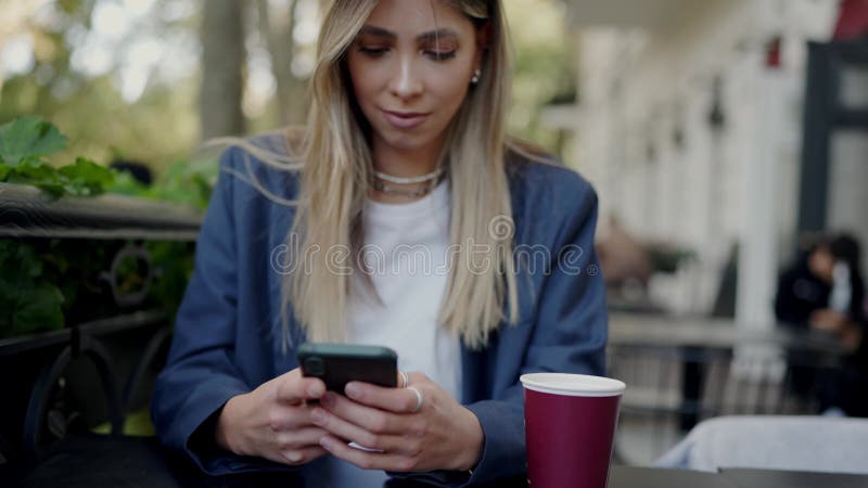 Young woman chats on smartphone in street cafe with cup of coffee on table