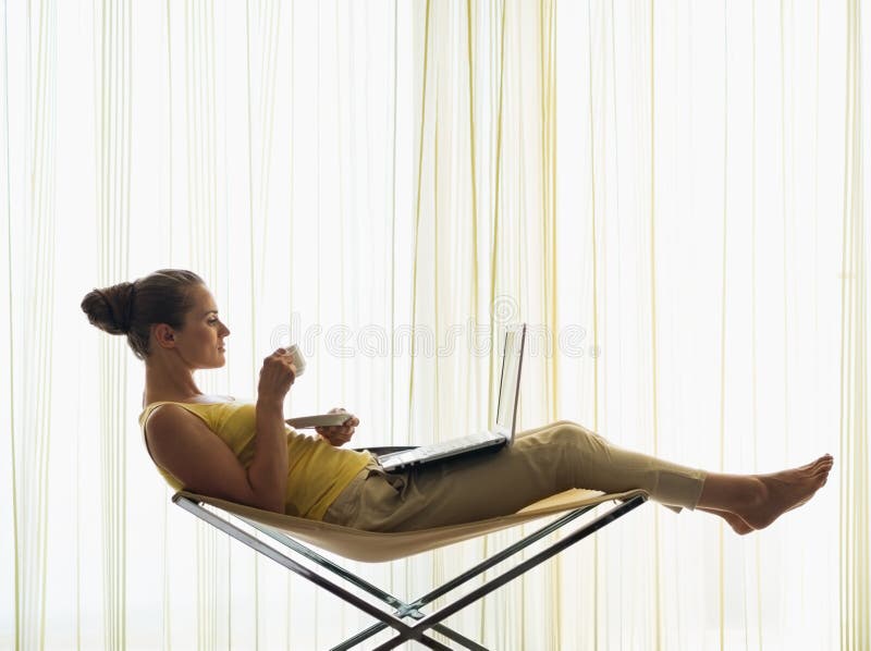 Young woman sitting on modern chair with coffee cup and using laptop. Young woman sitting on modern chair with coffee cup and using laptop