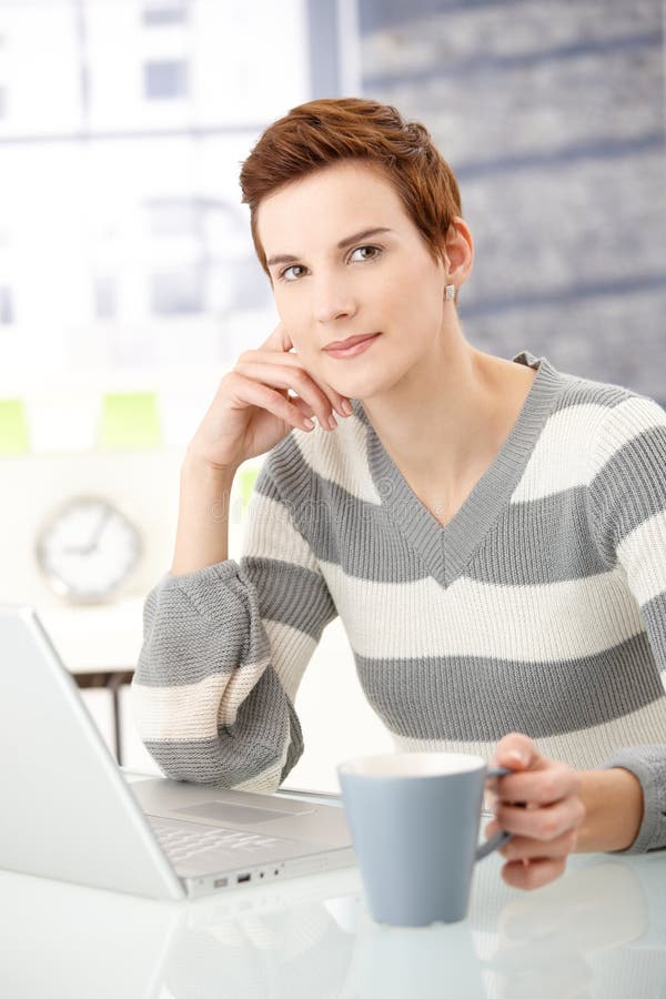 Portrait of young woman sitting at table with coffee mug and laptop computer, smiling at camera. Portrait of young woman sitting at table with coffee mug and laptop computer, smiling at camera.