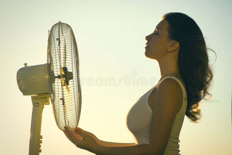 Young Woman cooling herself during hot weather in front of fan on a blue sunset sky background