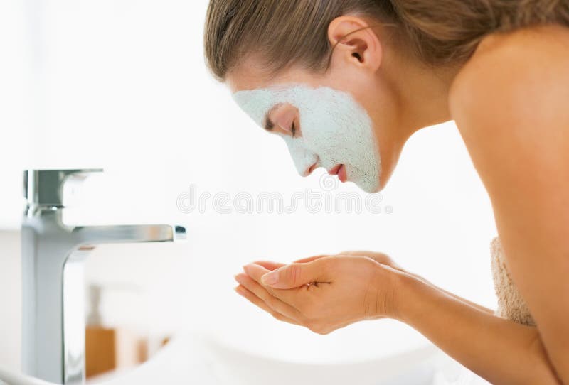 Young woman with cosmetic mask on face washing in bathroom