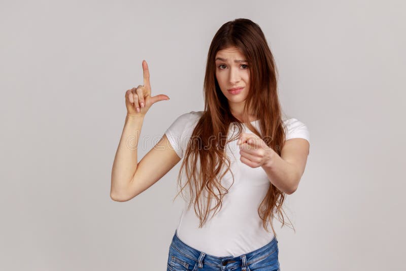 Young woman with dark hair looking at the camera, pointing at camera and showing loser gesture.