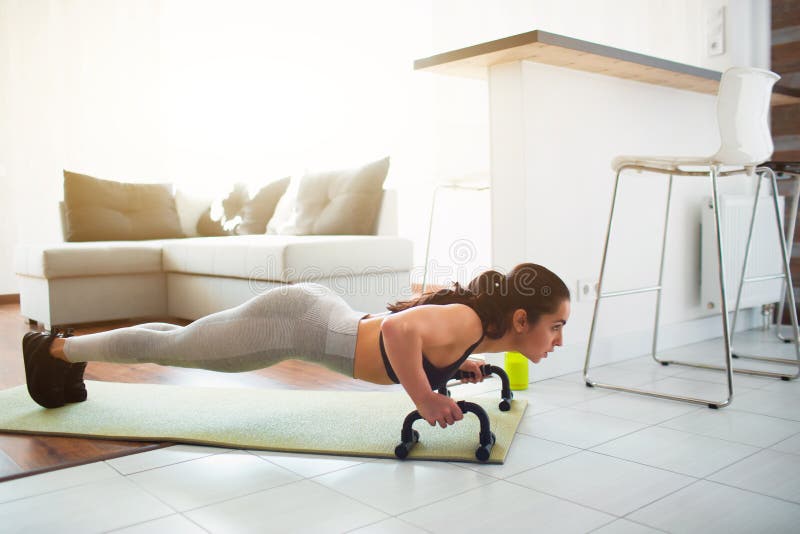 Young woman doing sport workout in room during quarantine. Fitness model stand in plank position using push up stand