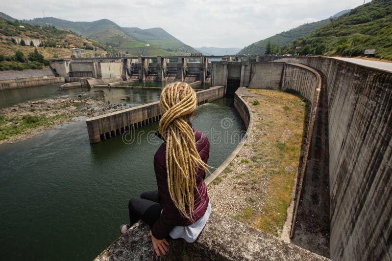 Young woman with dreadlocks sitting near a power plant in the mountains.