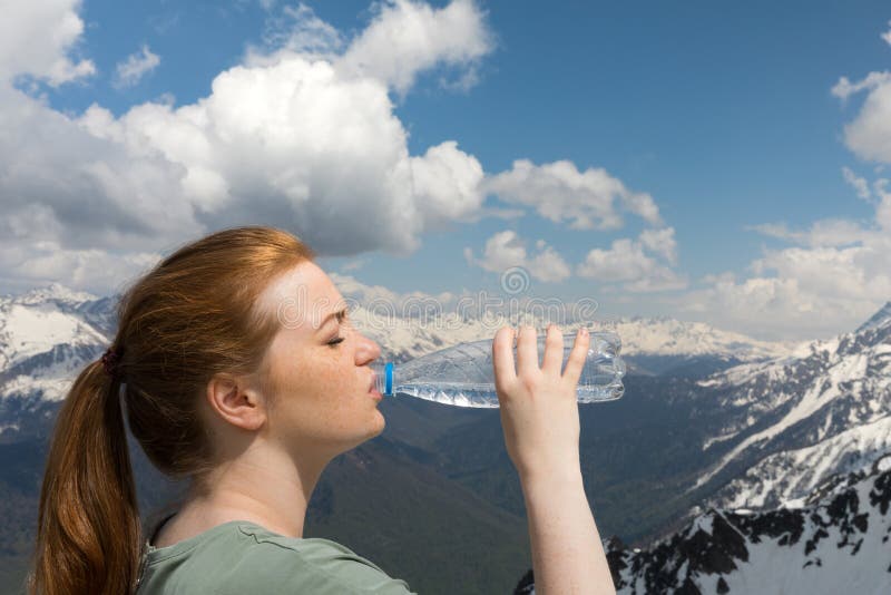 Young woman drink water from plastic bottle in the mountains on the snow peaks background