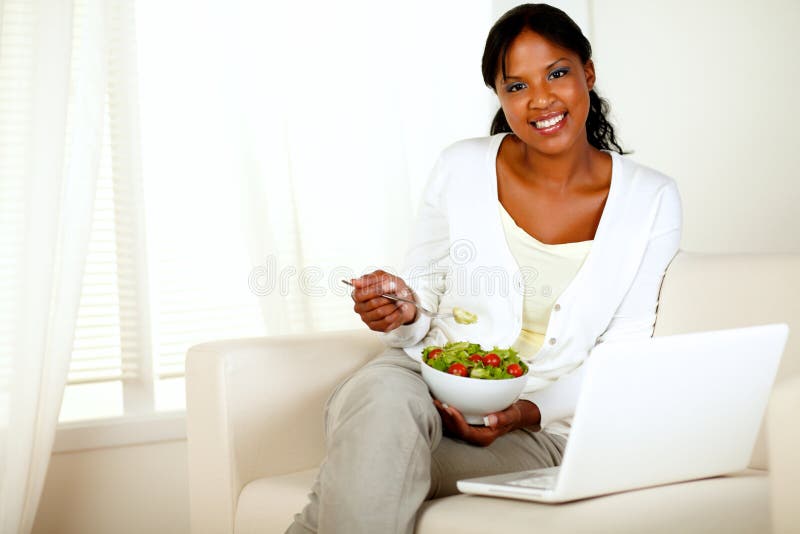 Portrait of a young woman eating healthy salad and looking at you while is sitting on couch in front of her laptop. with copyspace. Portrait of a young woman eating healthy salad and looking at you while is sitting on couch in front of her laptop. with copyspace