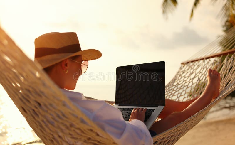 Back view of young woman, successful female freelancer using laptop while lying in hammock on the tropical beach at sunset, working remotely during summer vacation. Distance work concept. Back view of young woman, successful female freelancer using laptop while lying in hammock on the tropical beach at sunset, working remotely during summer vacation. Distance work concept