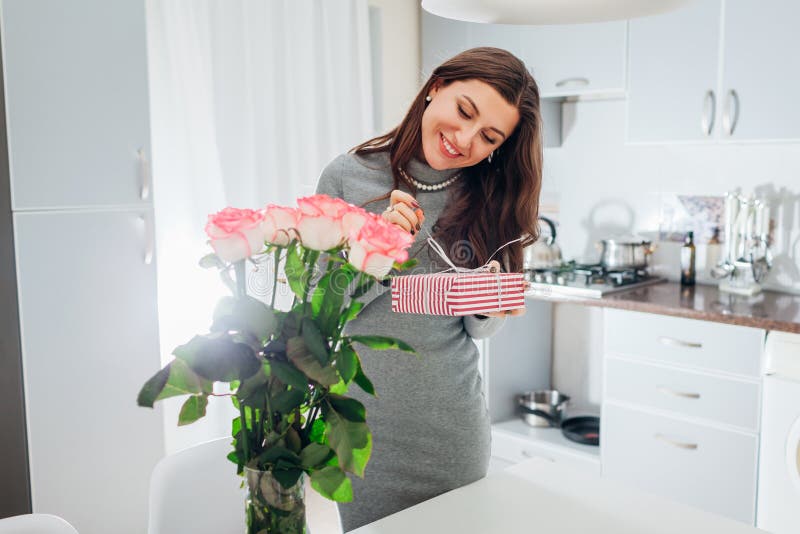 Young woman found gift box and bouquet of roses on kitchen. Happy smiling girl opening present stock photo Young woman found gift box and bouquet of roses on kitchen. Happy smiling girl opening present stock photo